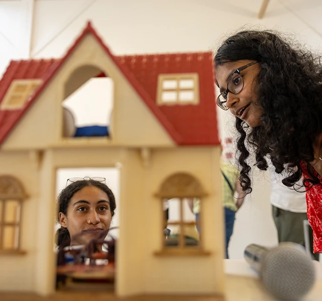 Two Knox students look at a dollhouse, one student is looking from above, while the other is peering in through a window.