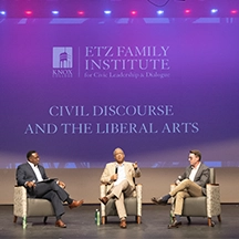 Three men in a discussion on a stage with the words, "Civic Discourse and the Liberal Arts,' on a screen behind them.