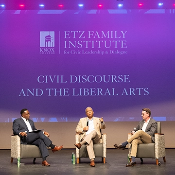 Three men in discussion on a stage with a screen behind them that states "Civlc Discourse and the Liberal Arts."