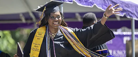 A Knox student wearing a mortar board, Commencement robe, and a variety of sashes waves as she walks across the Commencement stage 