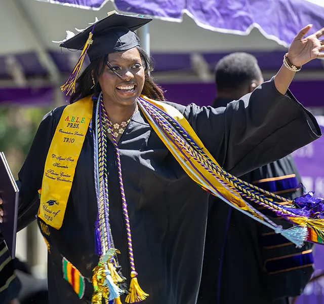 A Knox student wearing a mortar board, Commencement robe, and a variety of sashes waves as she walks across the Commencement stage 