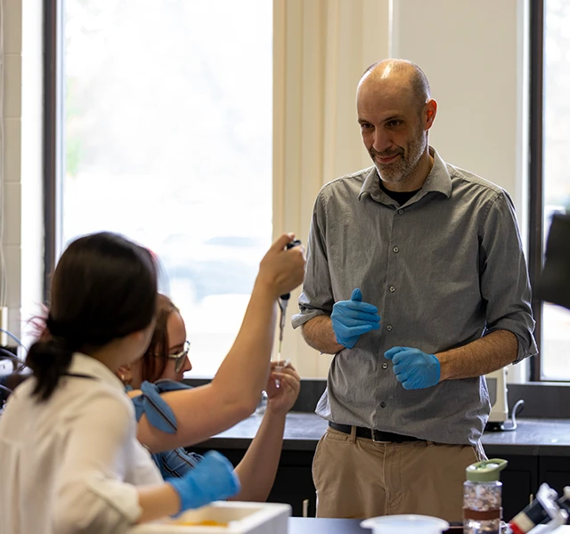 A Knox college science professor stands in front of two students. The professor is wearing blue surgical gloves, and one student is holding a pipette.